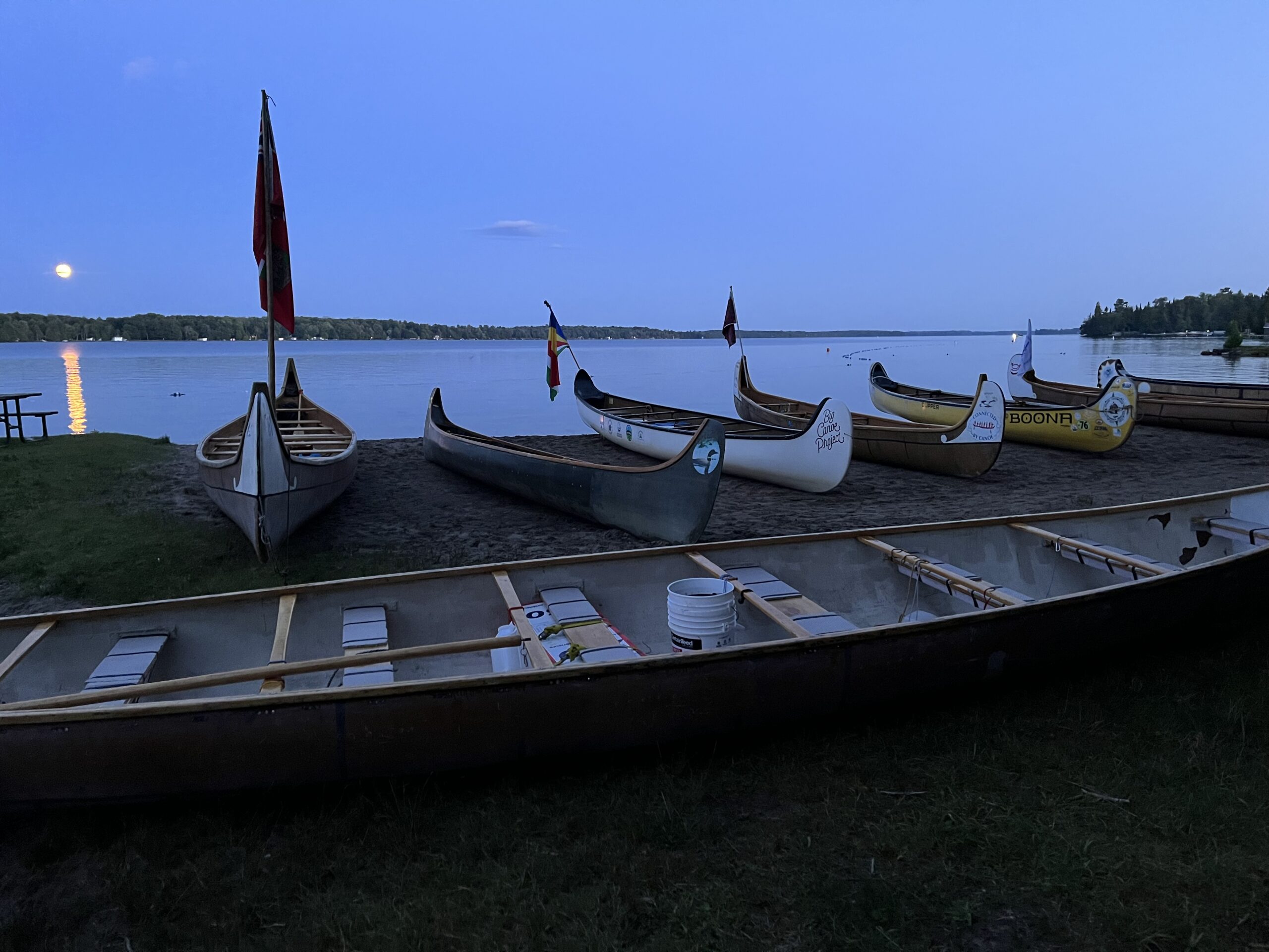 Fleet of voyageur canoes resting on shore at Balsam Lake at dusk with a full moon rising over the water