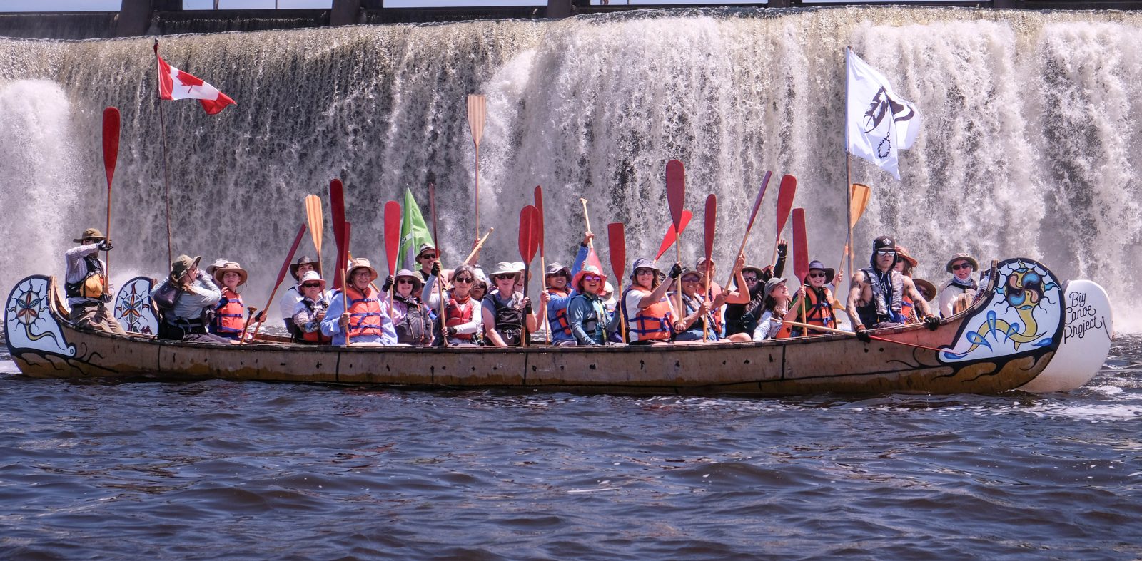 Paddlers in a painted Big Canoe Project voyageur canoe raise their paddles in front of Rideau Falls on the Ottawa River, flying Canadian and Algonquin flags