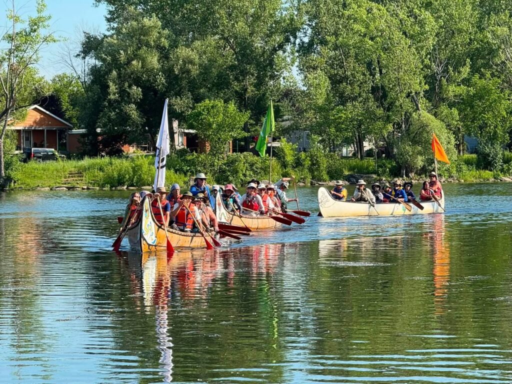 Three big voyageur canoes with flags paddling along a calm tree-lined stretch of the Ottawa River